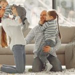 Cheerful mother kissing little boy while father hugging adorable daughter when family sitting on floor in rainy day against window